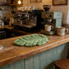 A green banana leaf absorbent hand towel resting on a wooden counter near coffee machines.