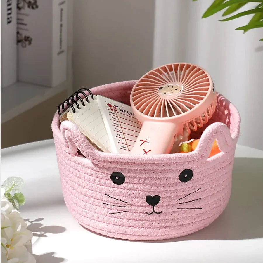 A pink woven cotton rope storage basket shaped like a cat face holds a small pink fan.