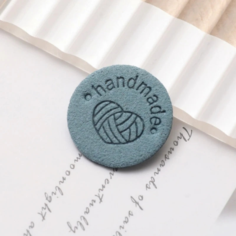 A round, teal-colored PU leather label with the word handmade embossed on it, resting on a piece of white paper.