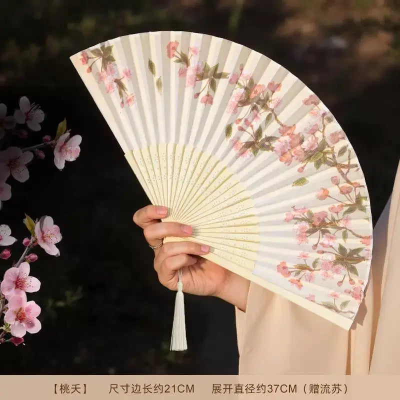 A pale pink and green floral patterned bamboo and silk folding fan being held by a person.