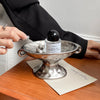 A vintage ceramic soap dish with an elevated stand holds a glass bottle of perfume on a wooden table.