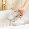 A hand is pouring water into a white double-layer draining soap dish in a bathtub.