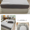 A fitted sheet in solid gray fabric is shown folded and laid out on a bed, with a hand demonstrating its elastic band.