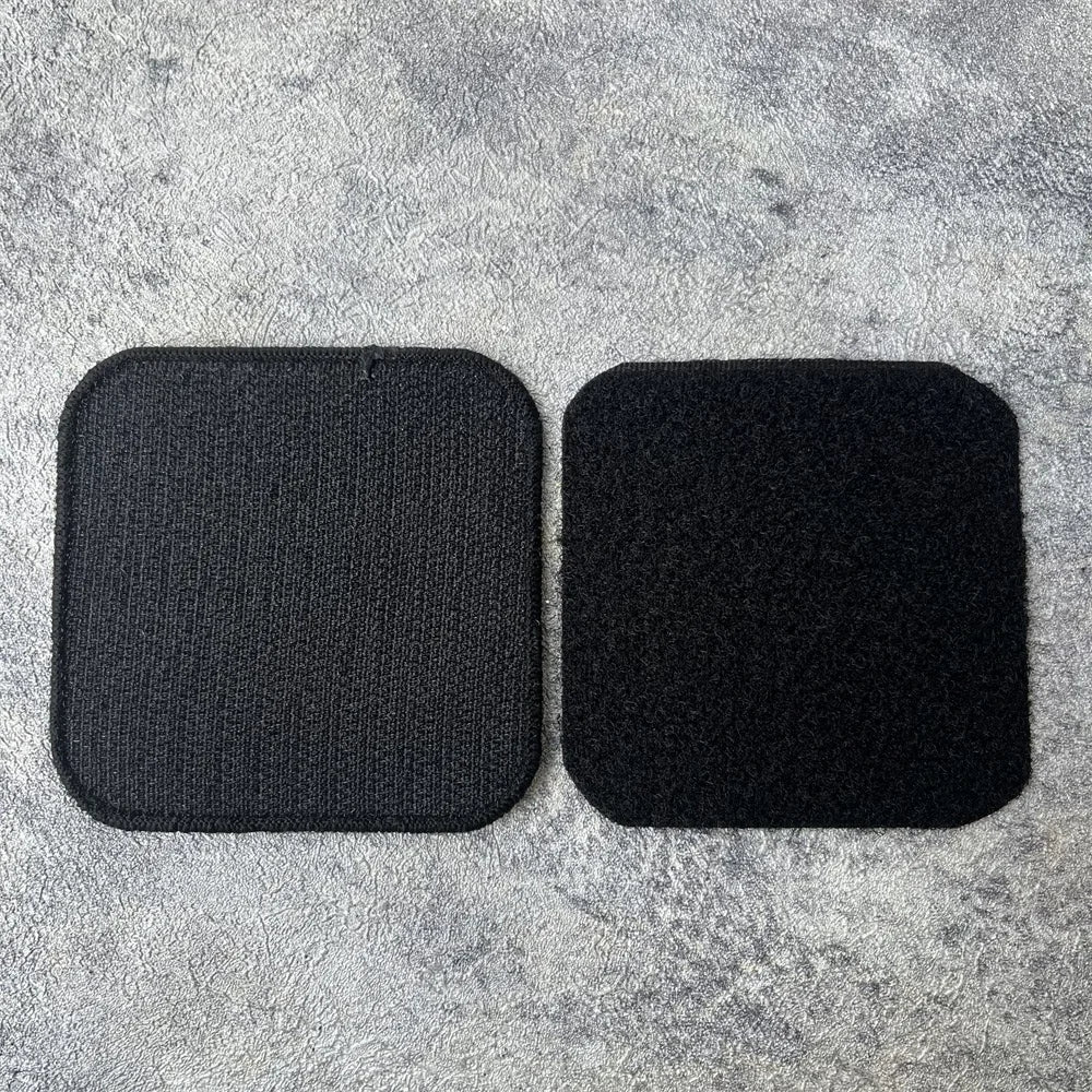 Two black embroidered tactical patches with hook and loop backing placed on a gray textured surface.