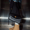 A person wearing a plaid shirt and dark pants holds a black leather square crossbody bag with a gold lock.