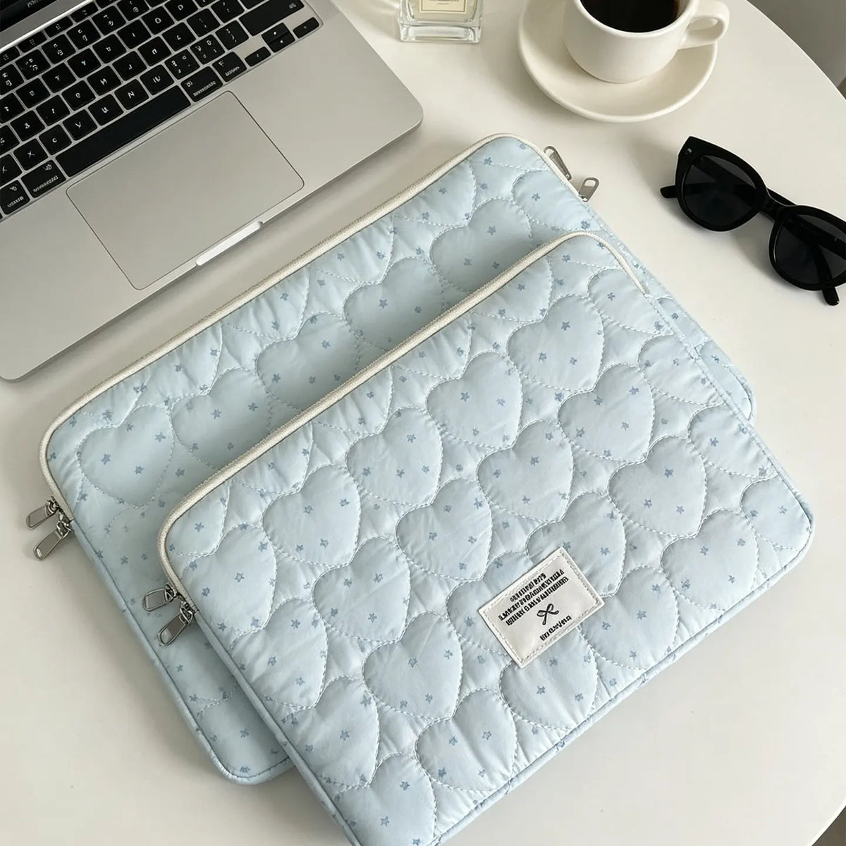Light blue quilted laptop sleeve resting on a white table next to a silver laptop and sunglasses.