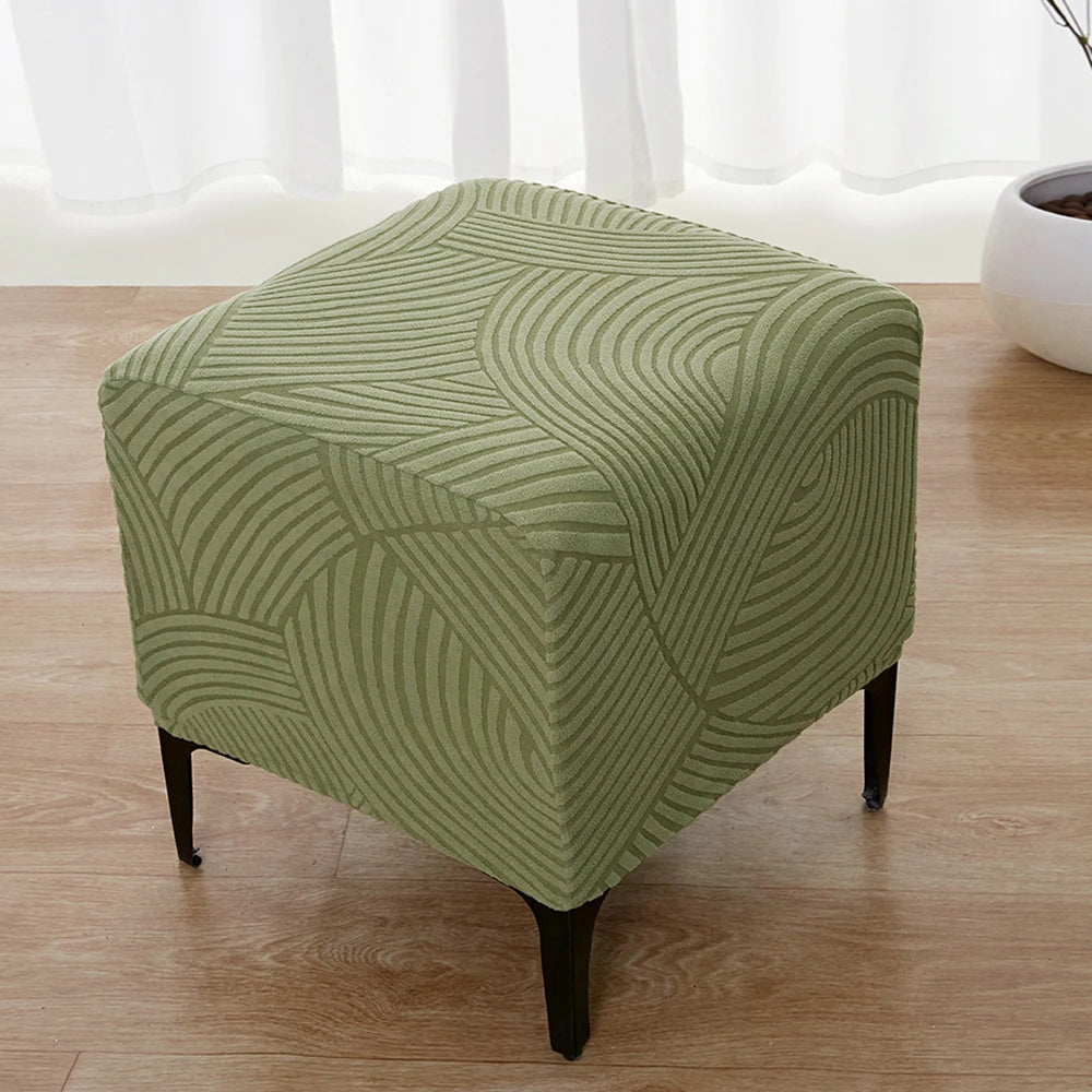 A square ottoman covered in a sage green geometric jacquard stretch fabric sitting on a wooden floor.