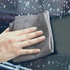 A hand holds a gray microfiber cleaning cloth against a wet car window.