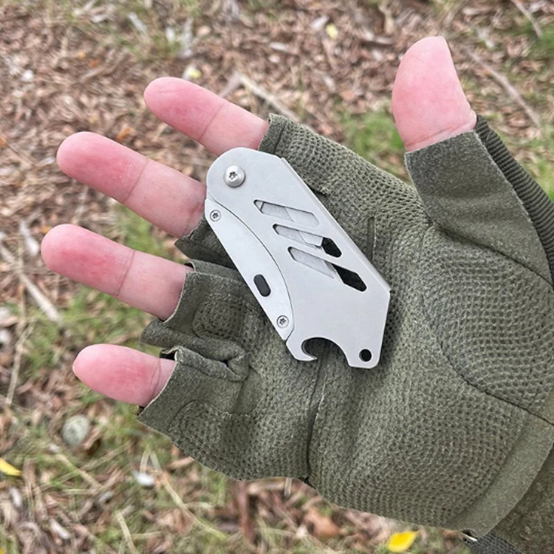 A hand holds a stainless steel folding utility knife with five replaceable blades against a green fabric glove outdoors.