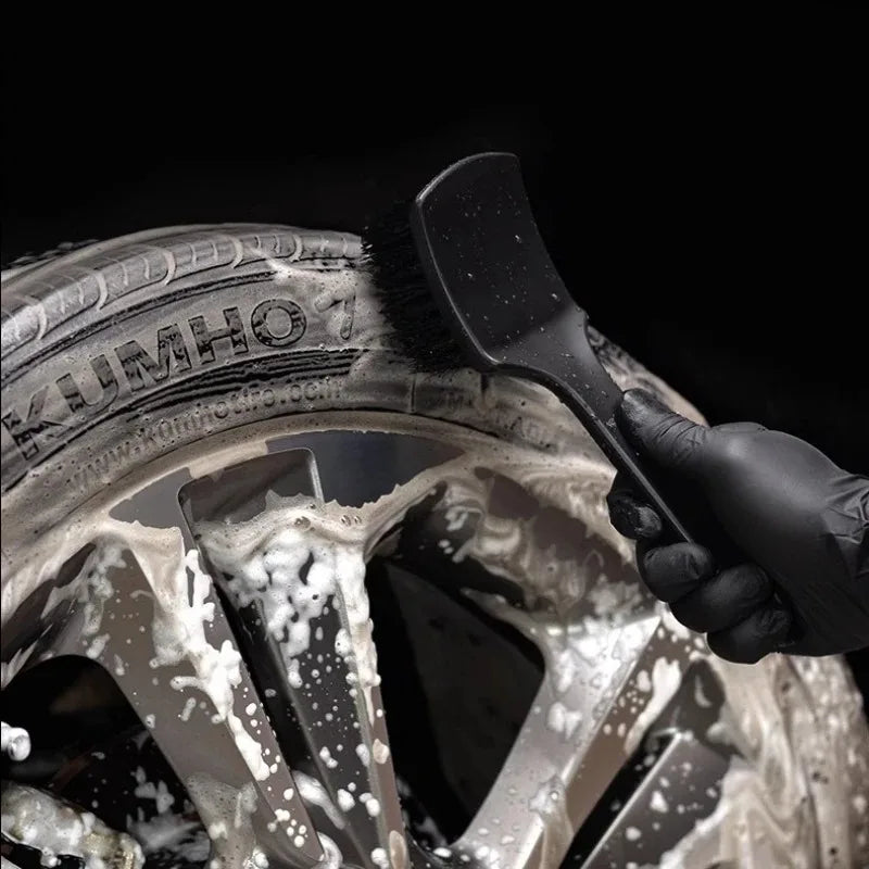 A hand uses a black brush to clean white foam from a silver alloy wheel rim.