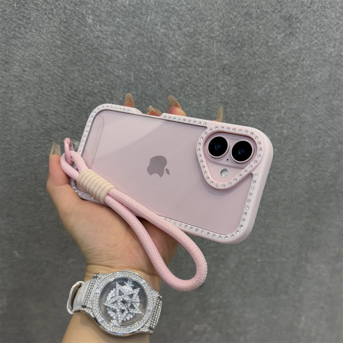 Una mano sostiene una funda rosa para iPhone con pulsera y un reloj transparente de diamantes.