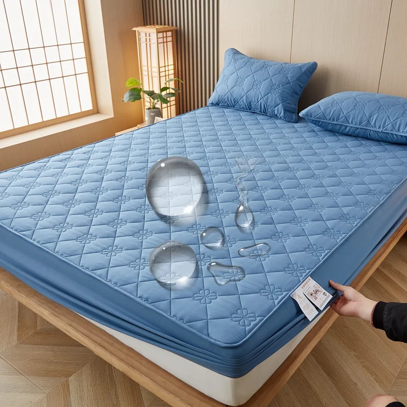 A blue quilted mattress protector is displayed on a wooden bed frame with pillows and a window in the background.