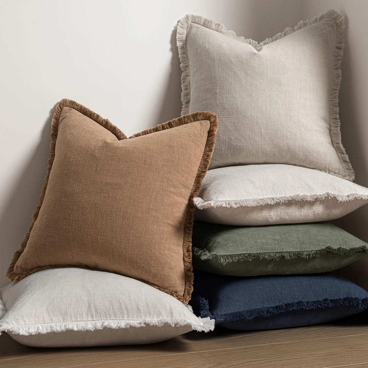 A stack of beige and olive green throw pillows with natural linen texture and fringe on a wooden surface.