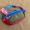 A purple and green unisex water-resistant crossbody chest bag with a pink tag resting on a brown textured surface.