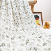 A white muslin baby swaddle blanket with a green leaf pattern draped on a surface.