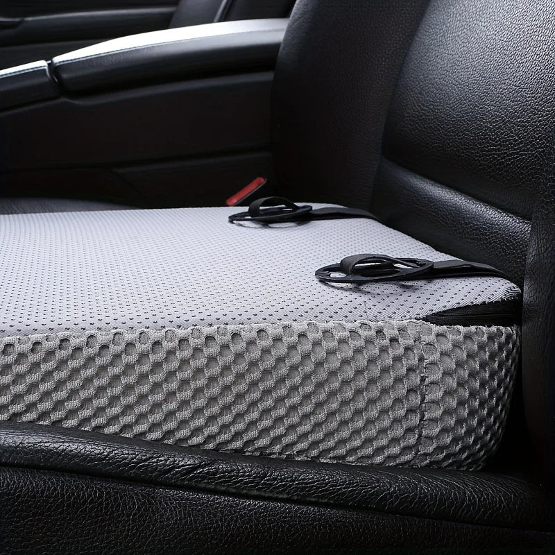 A gray ergonomic car seat cushion with a textured surface and black straps is placed on a black leather seat.
