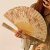 A hand holds a bamboo and silk folding fan with a floral design, featuring pale green and pink tones.