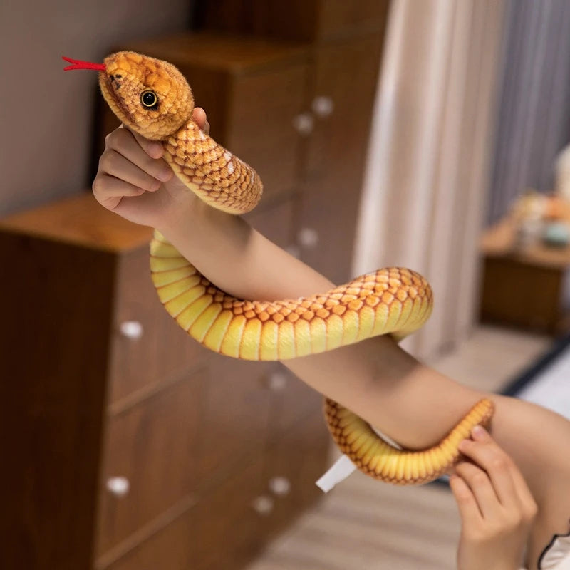 A hand is holding a lifelike, soft yellow snake plush toy with a red forked tongue.