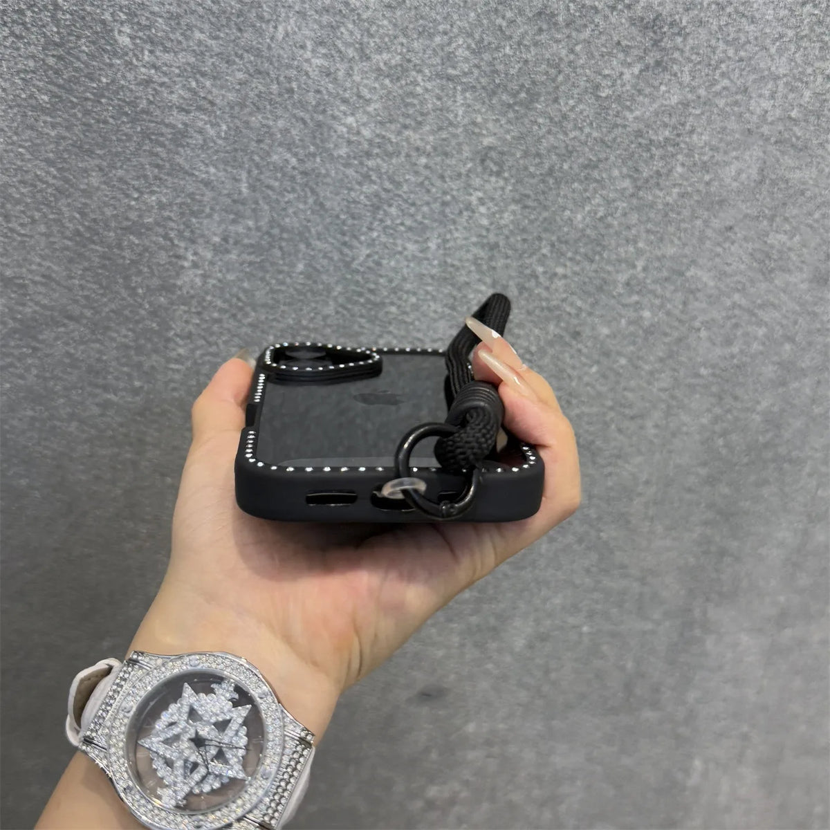 Una mano sostiene una funda de teléfono negra con correa de muñeca contra un fondo gris.
