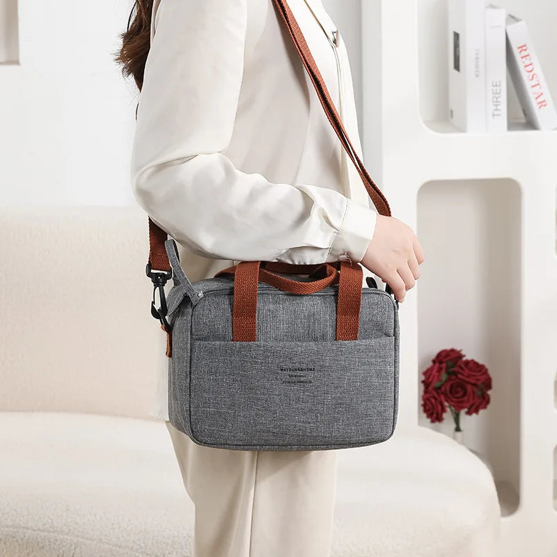 A woman holds a gray compact thermal lunch tote bag with brown leather straps in a bright indoor setting.