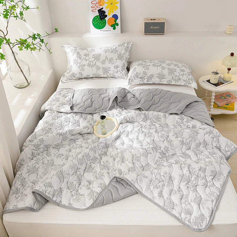 A white quilted blanket with a gray pattern is displayed on a bed, next to a plant and decorative items.