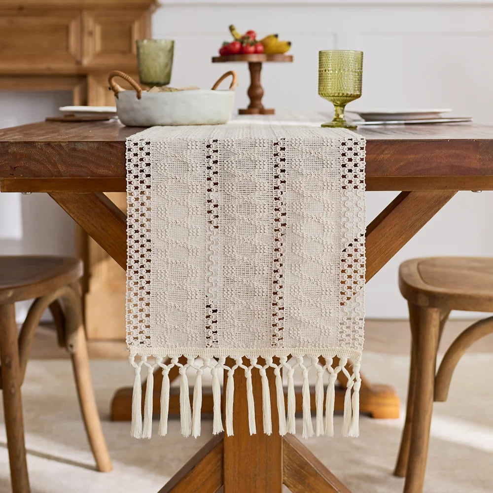 A beige hand-woven boho table runner with tassels is displayed on a wooden table.