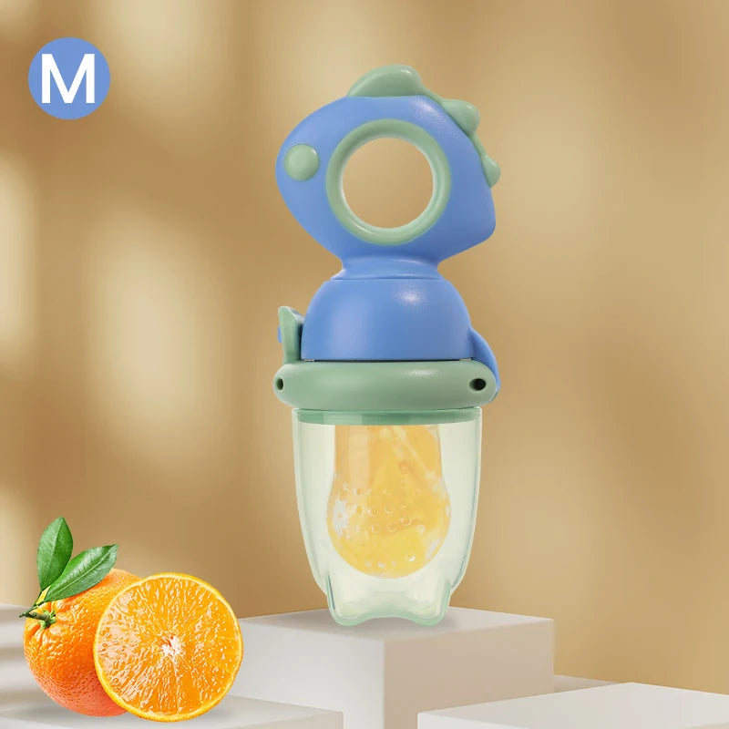 A blue and green rotating silicone baby fruit feeder with a dinosaur design, positioned next to an orange.