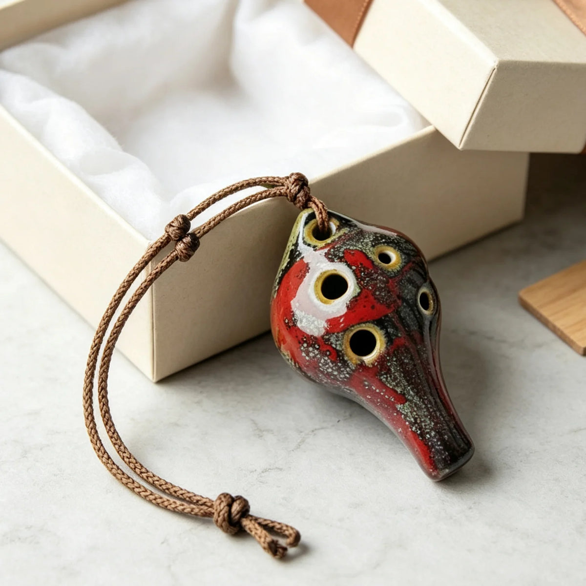 Handcrafted ceramic pendant ocarina with six holes, featuring red and white glaze, resting in a light box.