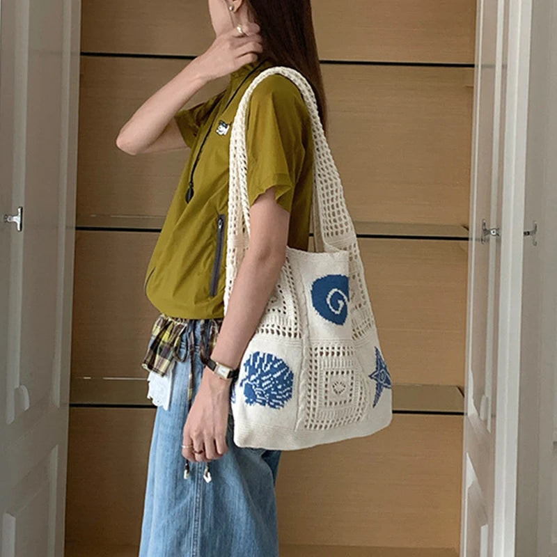 A woman wearing a olive green shirt and denim overalls holds a white handwoven knit tote bag with blue circular designs.