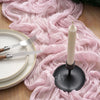 A pink crinkle gauze table runner flows over a wooden surface with a white candle and silverware.