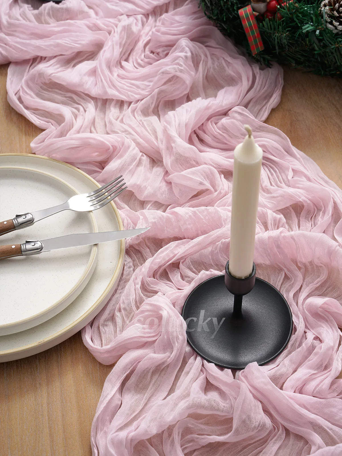 A pink crinkle gauze table runner flows over a wooden surface with a white candle and silverware.