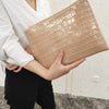 A woman holds a beige crocodile-embossed PU leather envelope clutch against a light background.