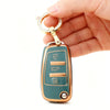 A hand holds a protective TPU key fob cover for an Audi flip key, featuring a gold-toned ring and a teal body.