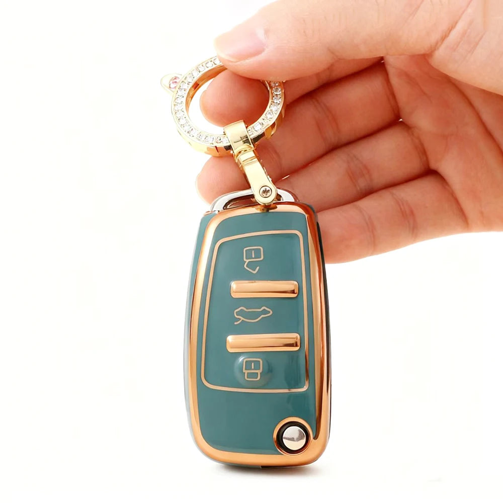 A hand holds a protective TPU key fob cover for an Audi flip key, featuring a gold-toned ring and a teal body.