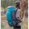 A person wearing a brown cap is carrying a teal and gray waterproof hiking backpack in an outdoor, wooded setting.