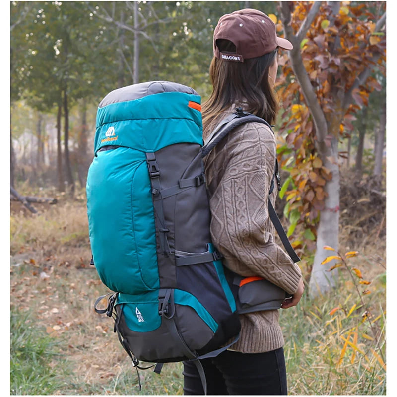 A person wearing a brown cap is carrying a teal and gray waterproof hiking backpack in an outdoor, wooded setting.