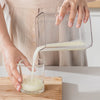 A hand pours yogurt from a stainless steel strainer into a glass bowl.