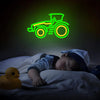 A neon sign shaped like a tractor in green and yellow glows against a dark background next to a sleeping baby.