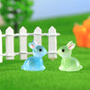 A set of cute resin miniature bunny figurines in blue and green colors placed on bright green grass behind a white picket fen