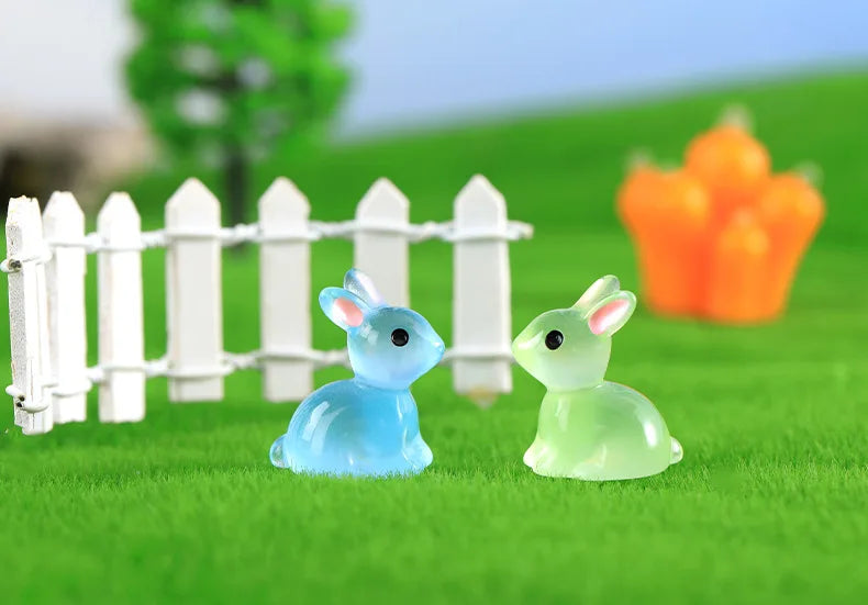 A set of cute resin miniature bunny figurines in blue and green colors placed on bright green grass behind a white picket fen