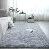 A fluffy gray silk-touch area rug placed on a light wooden floor in a bright room with sheer white curtains.
