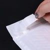 A hand is inserting a small, iridescent, teardrop-shaped object into a white padded bubble mailer.