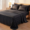 A black and gray striped satin bed sheet set is draped over a wooden bed with a lamp in the background.