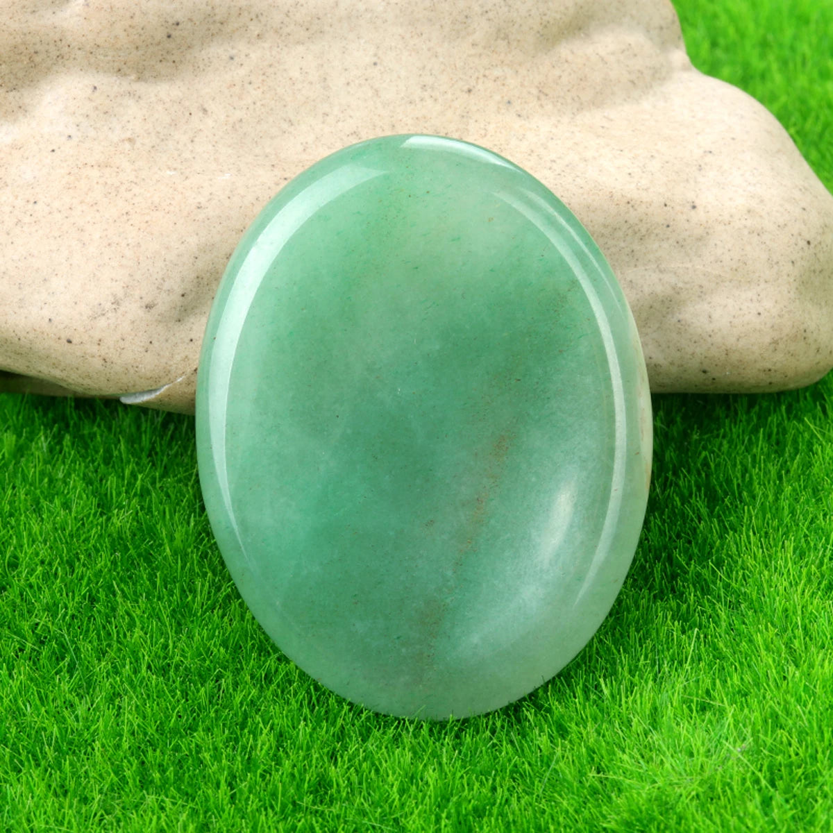 A smooth, oval green crystal stone resting on bright green grass, held by a hand.