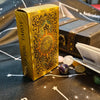 A gold foil tarot card deck box with intricate designs rests on a dark surface next to crystals and a guidebook.