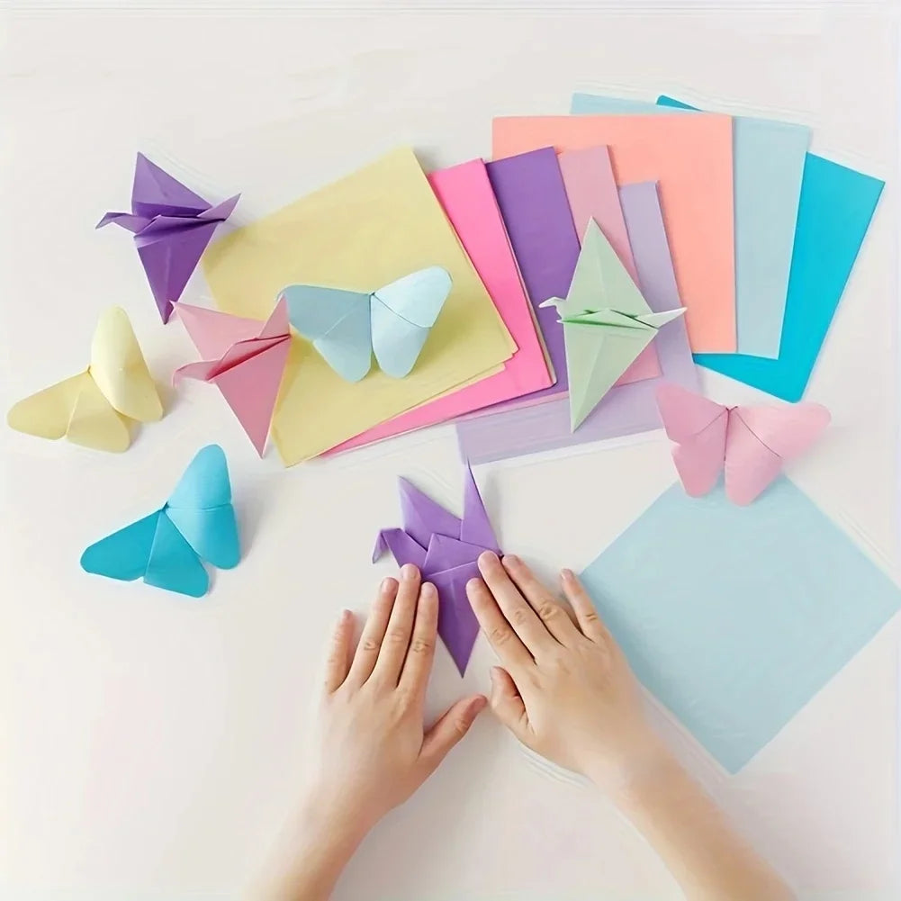 Hands are folding colorful origami paper into various shapes on a white surface.