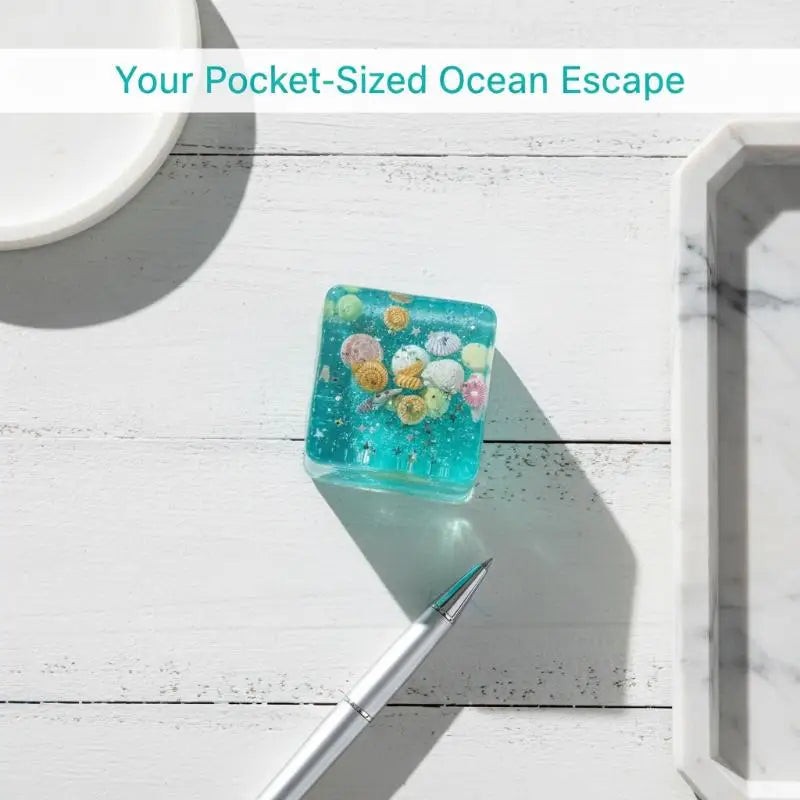 A small, translucent blue glass squeeze toy filled with colorful ocean-themed beads rests on a white wooden surface.