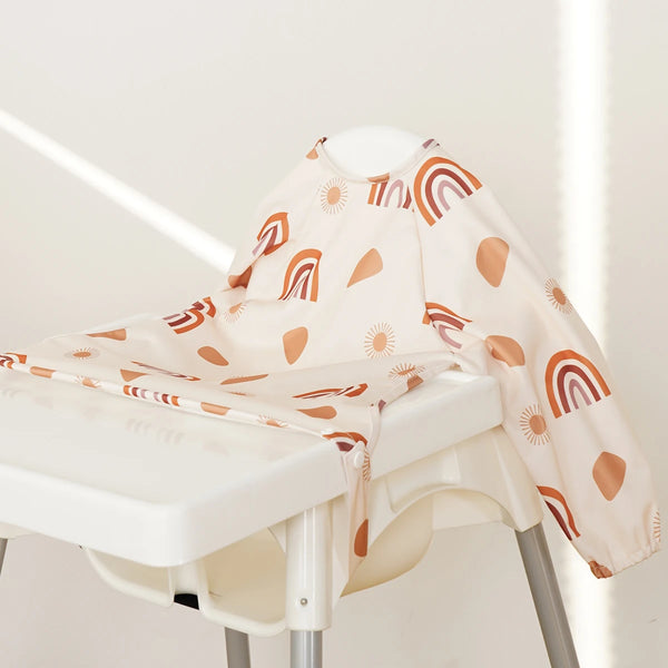 A beige baby feeding bib with long sleeves featuring a pattern of orange and brown rainbow shapes displayed on a white stool.