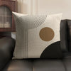 A beige linen cushion cover featuring black geometric lines and a circular tan accent on a dark sofa.