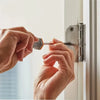 A person's hands are installing a door latch using a metal tool on a white window frame.
